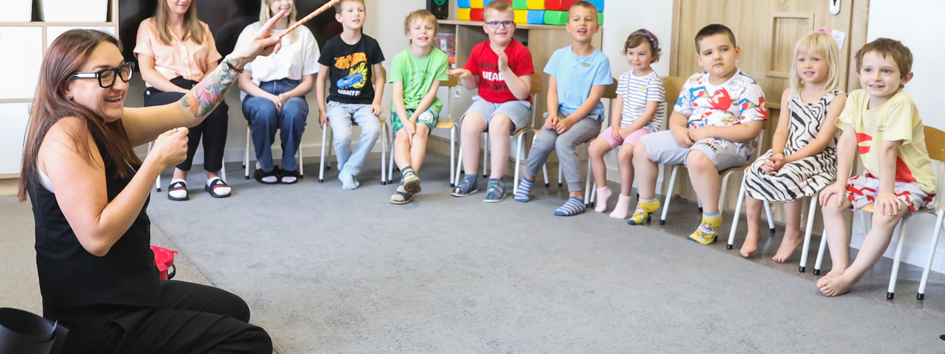 Kobieta siedzi na podłodze, wskazując kijem, podczas gdy grupa małych dzieci siedzi na krzesłach w klasie, uważnie ją obserwując.